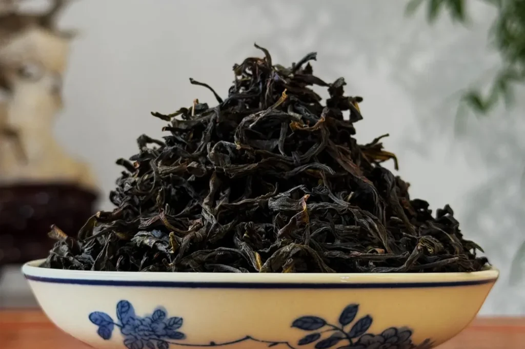 A blue-and-white porcelain bowl holds a tall mound of dark, dried loose-leaf tea in a close-up, with a softly blurred background.