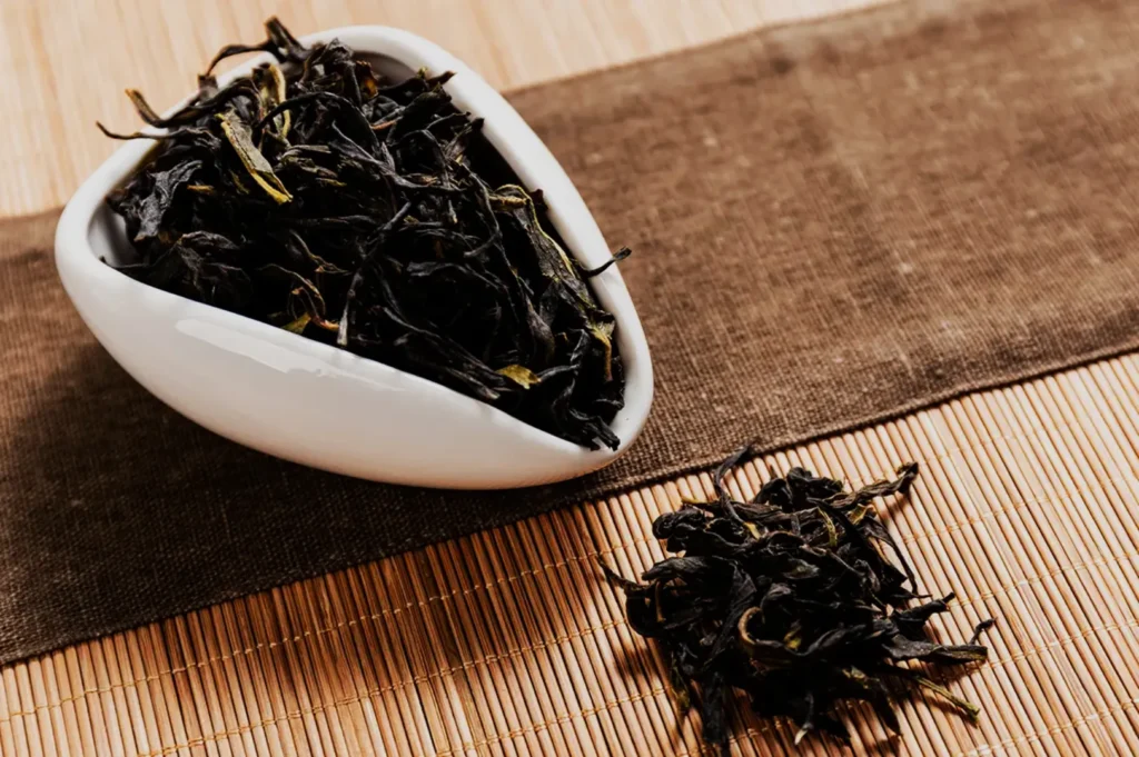 A white ceramic dish and loose pile hold dark, dried tea leaves on a bamboo mat and brown cloth, evoking a warm, rustic tea ritual.