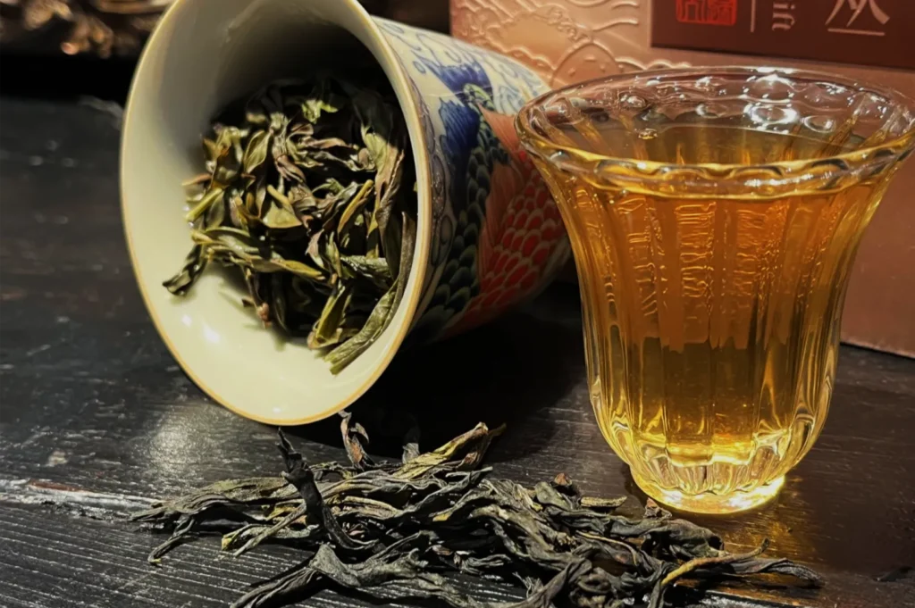 Loose tea leaves spill from a tipped blue-and-red patterned porcelain cup beside a glass of amber tea on a dark table, creating a warm, rustic mood.