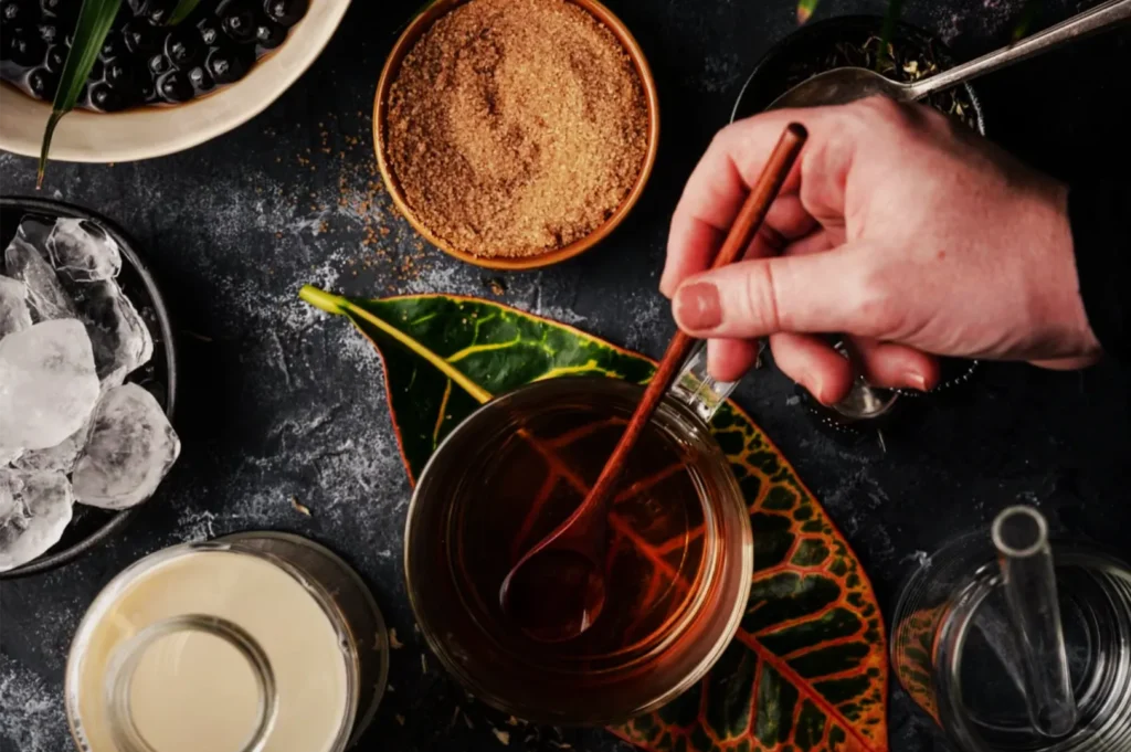 Overhead view of a hand stirring dark herbal tea in a glass, with berries, brown sugar, ice, and a pitcher on a leafy, dark tabletop.