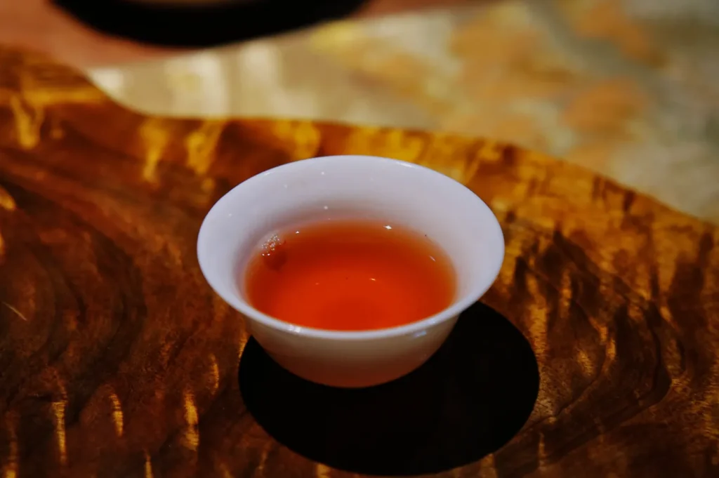 A small white ceramic cup filled with amber-red tea rests on a richly patterned wooden table, creating a warm, calm, intimate mood.