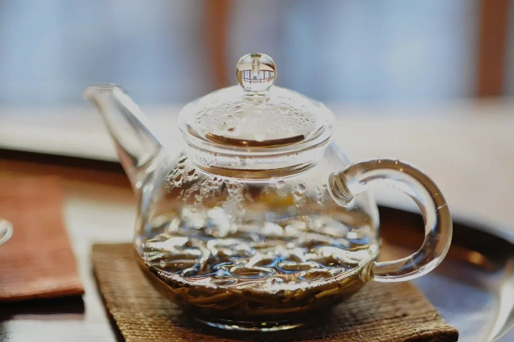Clear glass teapot with blooming tea steeping inside, resting on a woven coaster; warm natural light and soft focus create a calm, inviting mood.