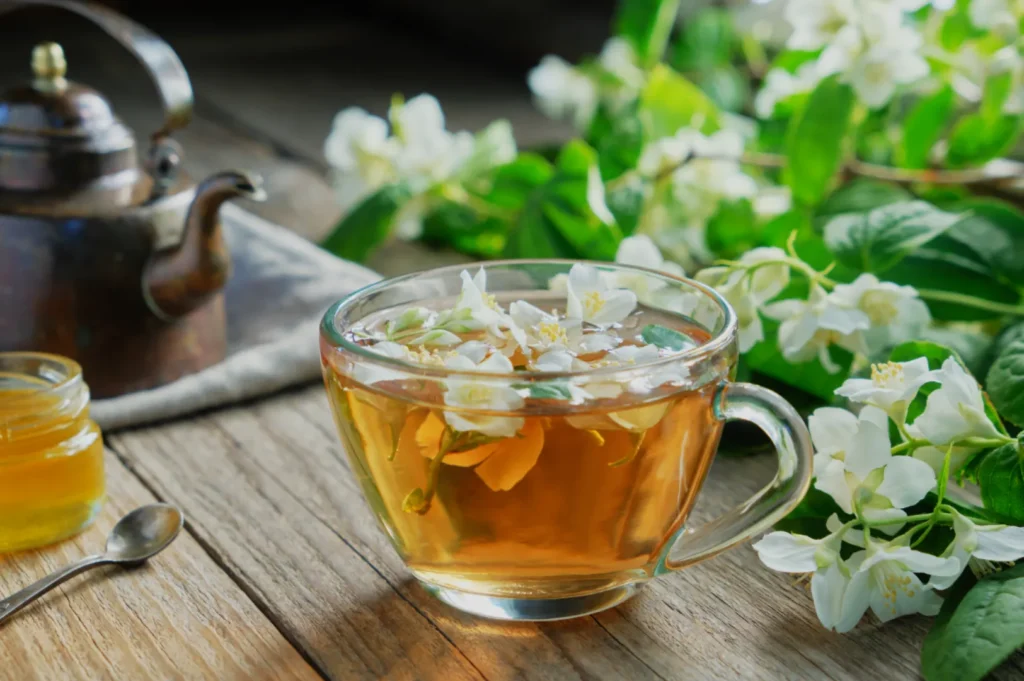 A glass cup of amber herbal tea topped with white blossoms sits on a rustic wooden table, beside a honey jar and kettle, surrounded by greenery.
