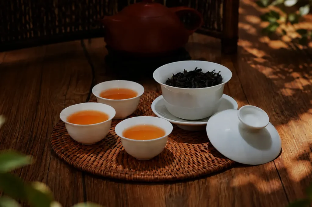 A serene tea service on a wooden table: three cups of amber tea, a bowl of loose dark leaves, and a clay teapot in warm, dappled light.