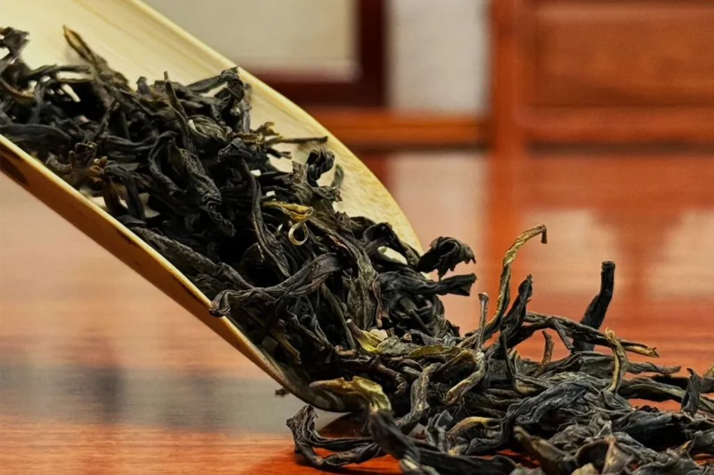 A wooden scoop pours dark, twisted loose-leaf tea onto a polished table, highlighting the dried leaves in a warm, quiet setting.