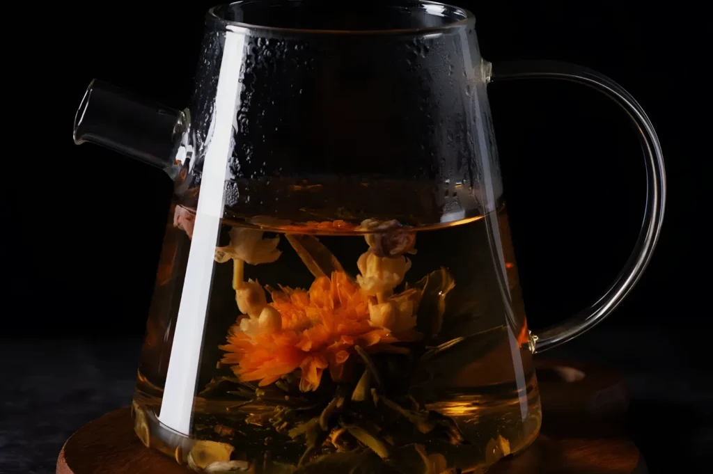 A clear glass teapot steeps orange and yellow flowers in amber tea against a dark background, creating a warm, elegant, calming mood.