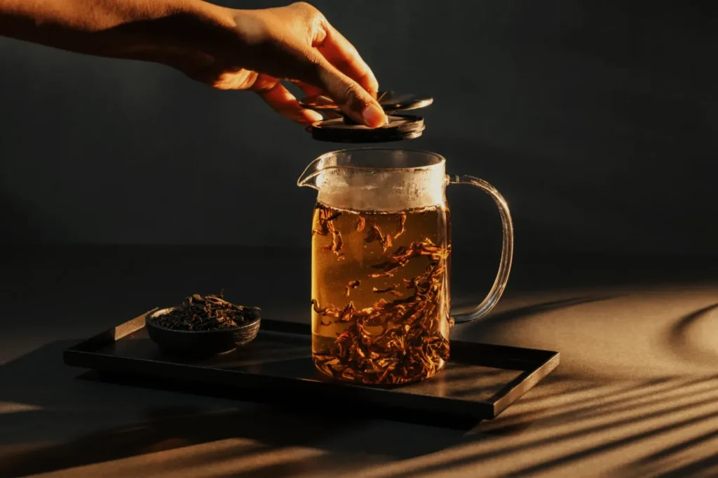 A hand places a lid on a glass teapot filled with amber herbal tea and floating leaves, beside a bowl of loose tea on a dark tray; warm, calm lighting.